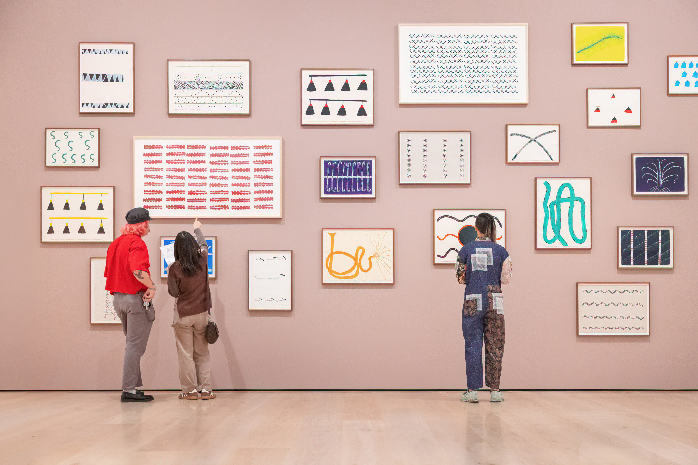 Three visitors, viewed from behind, stand in front of a pink gallery wall covered in a cluster of framed abstract artworks