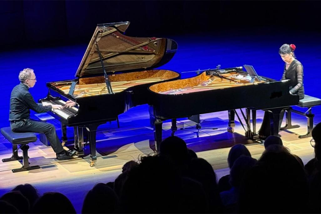 Gloria Cheng and Ralph van Raat perform on stage, lit by blue lighting. Both artists perform at their own pianos, positioned to be facing one another. The photo is taken from a high angle, showing the silhouettes of some audience members in the foreground.