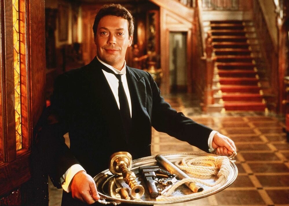 Tim Curry as Wadsworth in a tuxedo holds a tray of possible murder weapons in Clue.