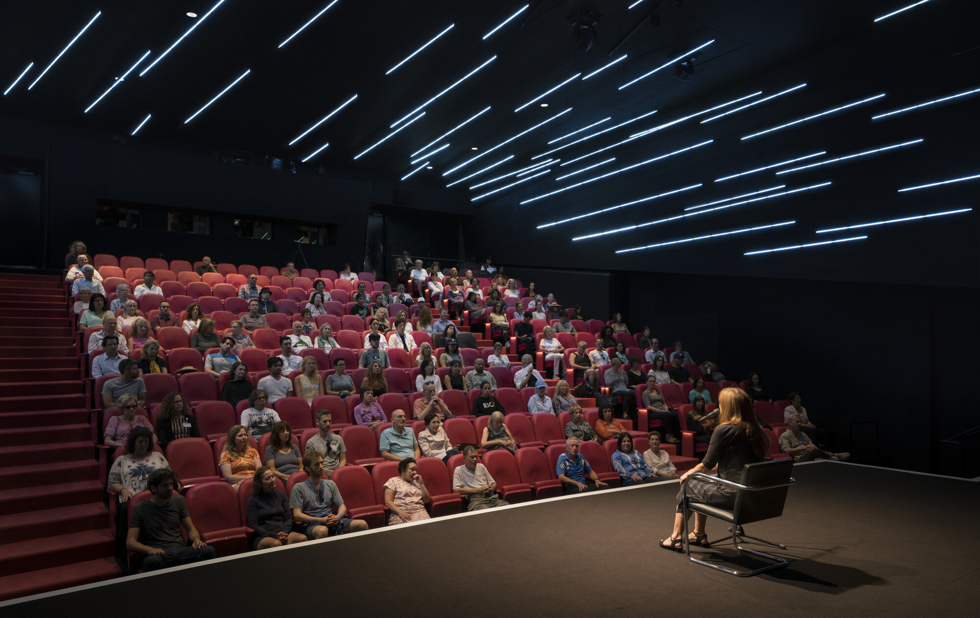 An audience sitting in a darkened theater, watching a woman on stage