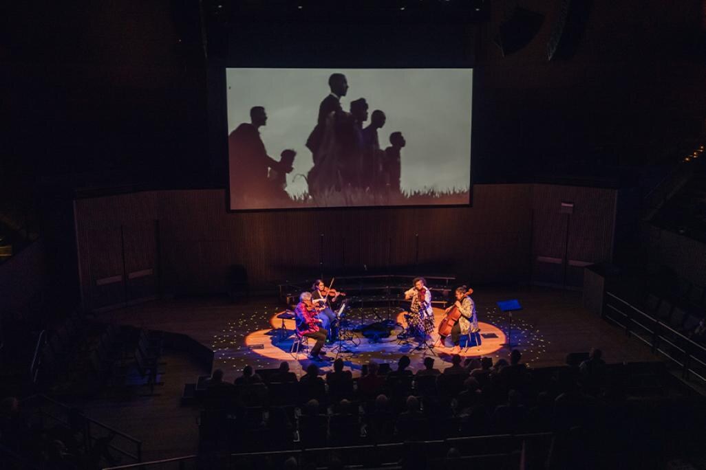 birds eye view of Kronos quartet members performing on stage amongst an audience with a projection in the background