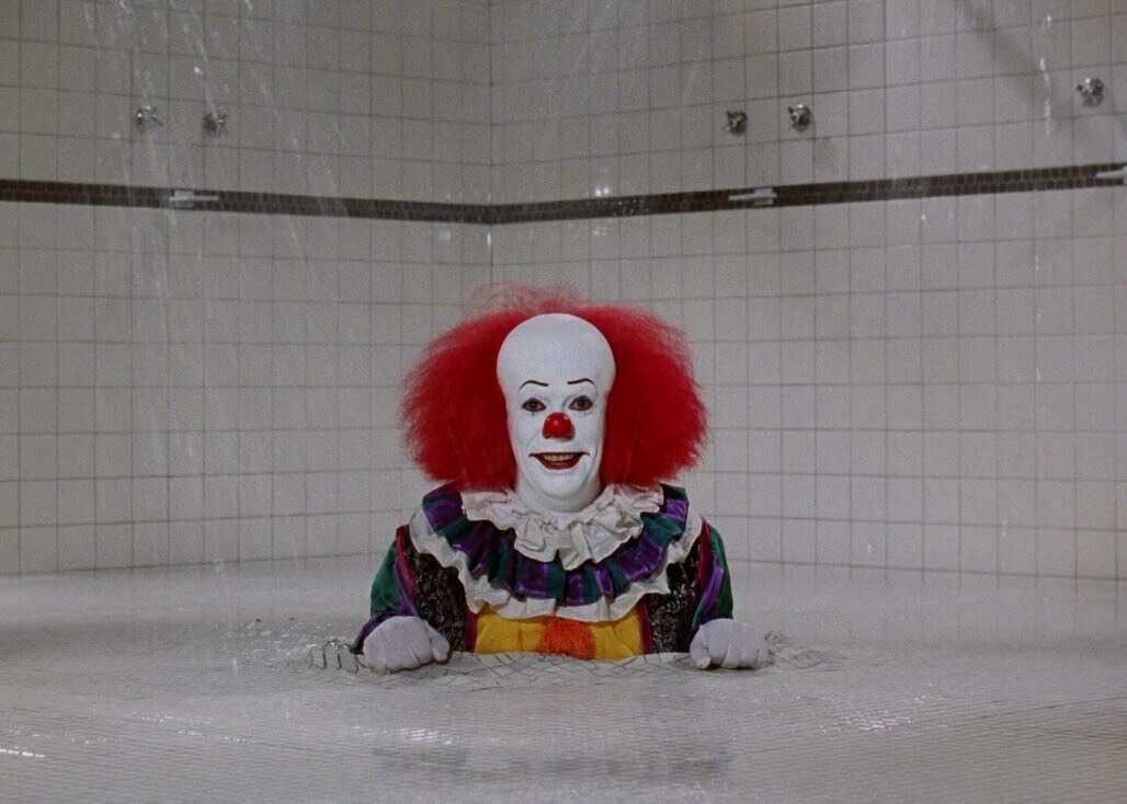 Tim Curry as Pennywise peers over the edge of a tiled shower in Stephen King's IT.